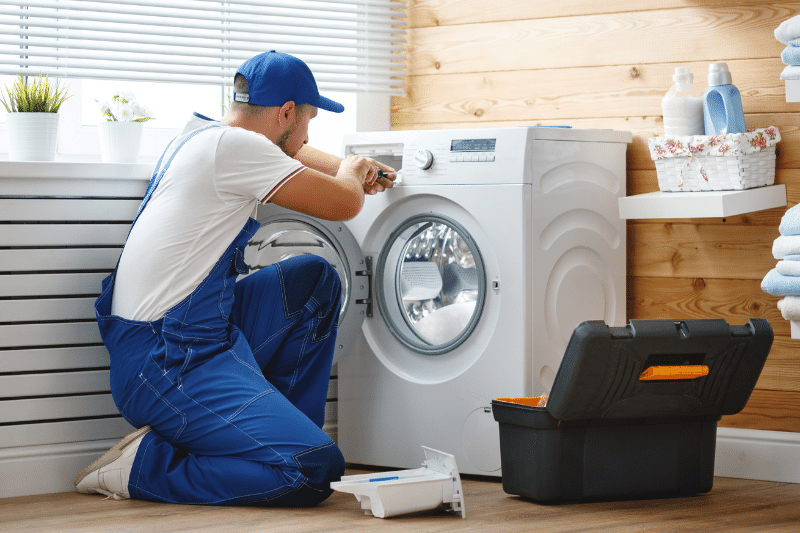 A repairman in blue overalls kneels in front of a washing machine, using a tool to fix it. An open toolbox sits nearby in a bright laundry room with folded towels and cleaning supplies.