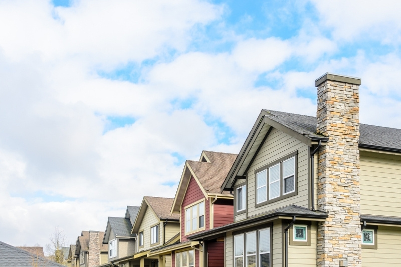 A row of modern suburban houses with varied siding colors and stone chimneys, seen under a partly cloudy blue sky.
