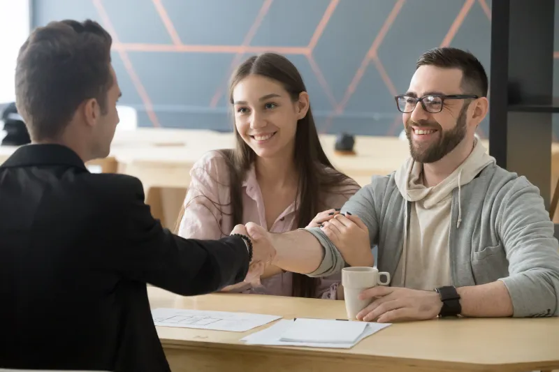 A man and woman smile while sitting at a desk, shaking hands with another man across from them. Papers and a coffee cup are on the table, suggesting a successful meeting or agreement.