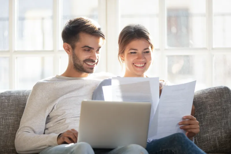 A smiling couple sits on a couch, looking at documents together. The man has a laptop on his lap, and both appear happy and relaxed, sitting in a bright room with large windows in the background.
